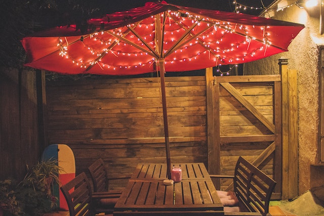 Brown wooden patio table with matching chairs and a red umbrella adorned with string lights