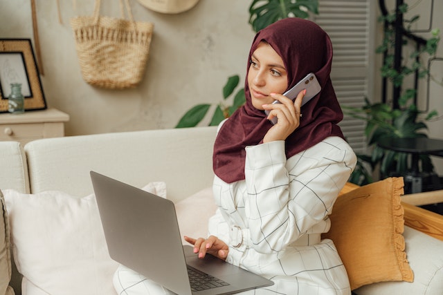 Person with a hijab sitting on a couch, holding a laptop, and making a phone call