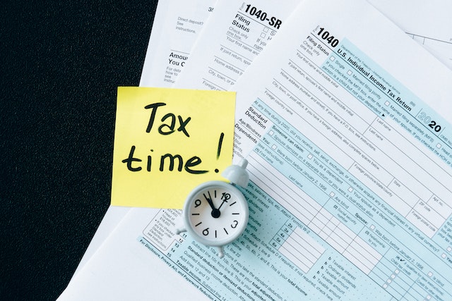 Yellow post-it that reads “Tax time!” next to a tiny clock and on top of U.S. tax documents