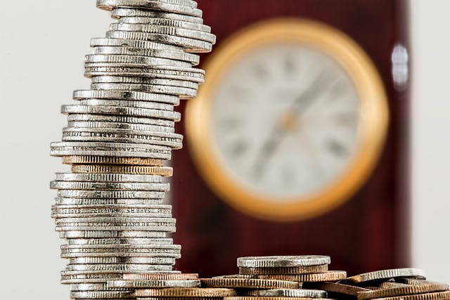 large-stack-of-coins-in-front-of-an-out-of-focus-clock