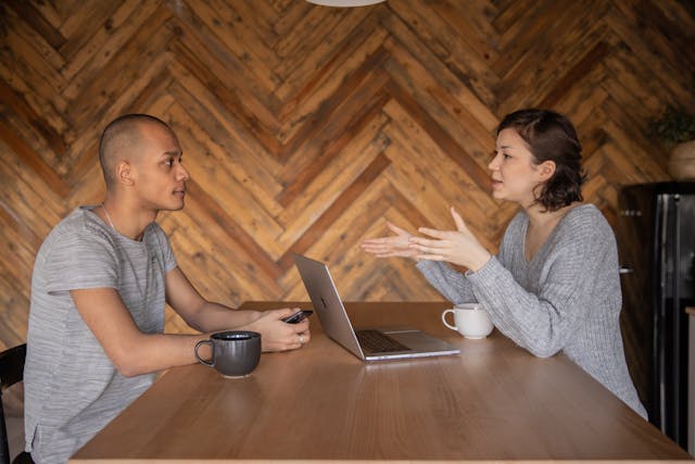  two-people-sitting-at-a-wooden-table-with-coffee-cups-and-a-laptop-in-front-of-one-person