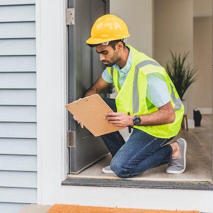 pexels-rodnae-productions-8293678 Person wearing a yellow hard hat and a yellow safety vest kneeling on a doorway and holding a clipboard